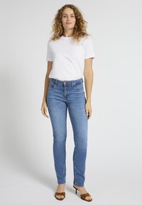 Kära juustega naine, kes kannab valget t-särki, siniseid kitsaid teksapükse ja pruune kontsaga sandaale, seisab ühetaolise heleda tausta ees.