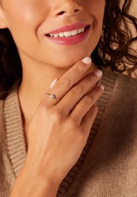 Zilveren ring met drie kleine sprankelende edelstenen, gedragen aan een hand met lichtgekleurde nagels, tegen een achtergrond van een gestructureerde beige breiselsweater.