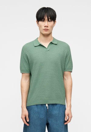 Νέος άνδρας με μαύρα μαλλιά φοράει ανοιχτό πράσινο πλεκτό πόλο με κοντά μανίκια και μεσαίου μπλε τζιν, στέκεται μπροστά σε ένα απλό φόντο.