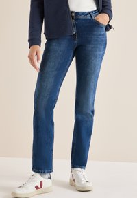 Jeans in denim blu scuro con taglio a gamba dritta, dotati di una chiusura a bottone classica e una leggera scoloritura, abbinati a sneakers bianche.