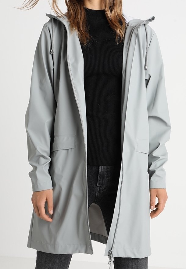 Personne portant un imperméable à capuche gris clair par-dessus un pull noir côtelé et un jean foncé, debout, les mains relâchées le long du corps.