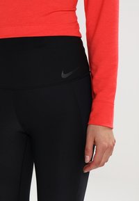 Czarne legginsy Nike o wysokim stanie z subtelnym logo, w połączeniu z dopasowaną, długą, czerwono-jasną bluzką wykonaną z miękkiego materiału.