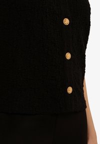 Zwarte sweater met textuur en vier gouden decoratieve knopen aan de zijkant. De stof heeft een gekreukt patroon, wat visuele interesse toevoegt.