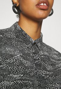 Svart och vit mönstrad skjorta med spetsig krage, texturerad tyg och abstrakta mönster, tillsammans med stora svarta örhängen.