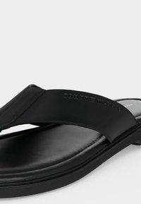 Μαύρη σαγιονάρα Tommy Hilfiger με επενδεδυμένο πάτο και φαρδιά λουράκι με ανάγλυφο λογότυπο της μάρκας σε απλό ανοιχτό φόντο.