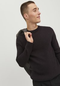 Maglione pullover viola scuro con polsini e orlo a coste, caratterizzato da un collo rotondo. Un blazer grigio appoggiato sulla spalla con motivo a quadri.