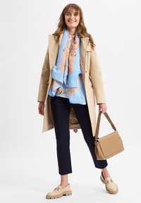 Lachende vrouw in een beige jas, met een blauwe gestreepte sjaal, witte blouse, zwarte broek, beige instappers en een beige handtas met een logoband.