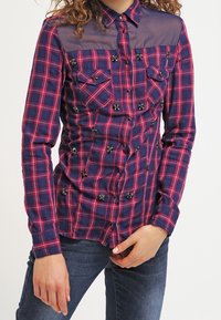 Chemise à carreaux bleu marine et rose avec un empiècement transparent, fermeture à boutons, deux poches poitrine et embellissements floraux décoratifs. Manches longues.