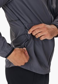 Grau leichtgewichtiges Jackett mit einer seitlichen Reißverschlusstasche, ausgestattet mit einem strukturierten Stoff und elastischen Bündchen. Hand zieht an der Öffnung der Tasche.