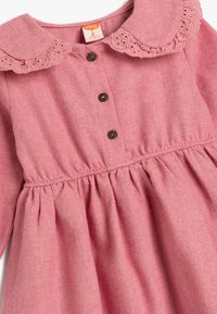 Rotes Wollkleid mit einem runden Spitzenkragen, fünf braunen Knöpfen und gerafftem Taillenbereich, das eine weiche Textur und ein klassisches Design aufweist.