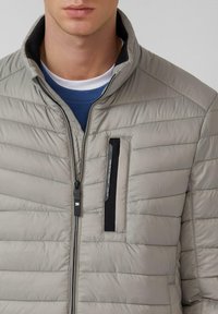 Lichtgrijs donsjack met een horizontaal doorgestikt patroon, hoge kraag, ritssluiting aan de voorkant en een borstzak met rits en zwarte accenten.