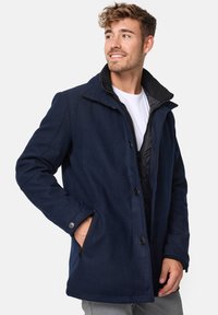 Manteau en mélange de laine bleu marine avec un col montant, fermeture à glissière et boutons, deux poches latérales, doublure intérieure texturée et poignets boutonnés.