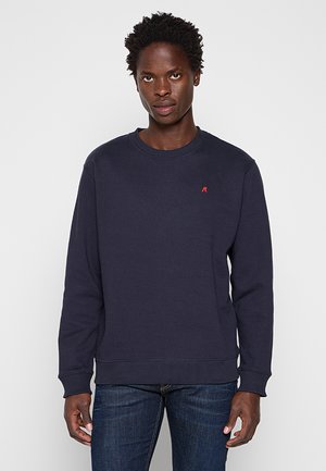 Mann trägt einen schlichten dunkelblauen Pullover mit kleinem rotem Logo auf der Brust und dunkelblaue Jeans, steht vor hellem Hintergrund.