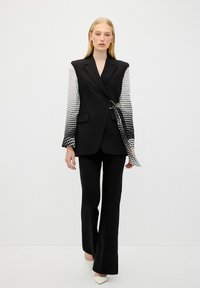 Blazer noir avec des manches à motifs, cintré à la taille avec un lien. Assorti à un pantalon noir évasé et des talons blancs pointus.