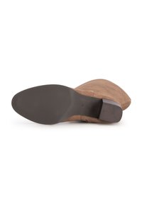 Botte en suède marron couchée sur le côté montrant sa semelle texturée noire et son talon bloc sur fond blanc.