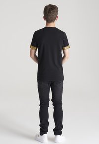 Camiseta negra con detalles amarillos en las mangas cortas, combinada con jeans negros ajustados y zapatillas blancas. Textura suave en general.