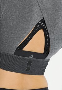 Das graue Sportoberteil zeichnet sich durch ein strukturiertes Design mit einem breiten elastischen Saum aus und verfügt über ein Ausschnittdetail, das eine schwarze gemusterte Unterschicht offenbart.