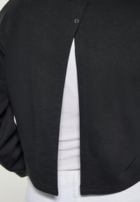 Chaqueta corta negra con un acabado texturizado, que presenta una abertura trasera asegurada por un broche plateado. Combinada con una camiseta blanca por debajo.