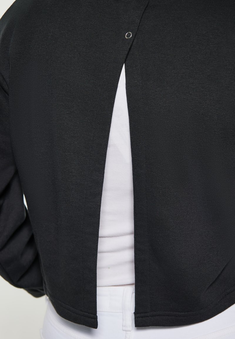 Chaqueta corta negra con un acabado texturizado, que presenta una abertura trasera asegurada por un broche plateado. Combinada con una camiseta blanca por debajo.