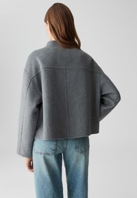 Vrouw met lang bruin haar, gekleed in een losse grijze wollen jas en lichtblauwe jeans, die met één hand op haar heup en met haar gezicht weg van de camera staat.