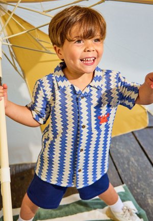 Jonge jongen lacht en houdt een gele paraplu vast, draagt een blauw-wit shirt met zigzag patroon en donkerblauwe korte broek.