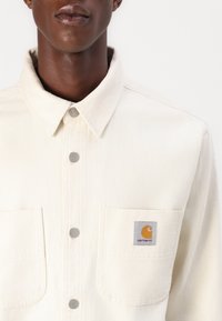 Világos bézs denim ing gombos elejével, két mellzsebbel és ezüst színű fémrészekkel. A bal zsebén Carhartt logó található.