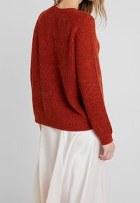 Pull en tricot couleur rouille avec texture côtelée, encolure ronde et coupe décontractée. Porté sur une jupe blanche en soie, mettant en valeur un effet de superposition.