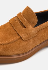 Mocasines de ante marrón con una textura suave, que presentan un sutil acento de correa y una suela de goma resistente. Diseño clásico con forma redondeada.