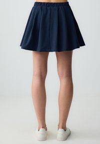 Marineblaue Faltenrock mit elastischem Bund, der oberhalb des Knies hängt, kombiniert mit weißen Sneakers. Glatte Stofftextur.
