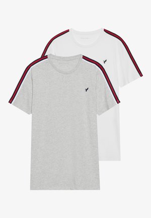 Deux t-shirts à col rond, l'un gris et l'autre blanc, présentant des rayures rouges et bleu marine contrastantes sur les épaules. Mélange de coton, manches courtes.