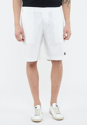 Uomo con pantaloncini bianchi fino al ginocchio, scarpe da ginnastica bianche e maglietta nera, in piedi con le mani rilassate ai lati.