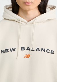 Beige Kapuzenpullover mit vorderer Kängurutasche, verstellbarem Kapuzenzug und großem "NEW BALANCE"-Druck in Schwarz und Orange. Aus weichem Baumwollmaterial.