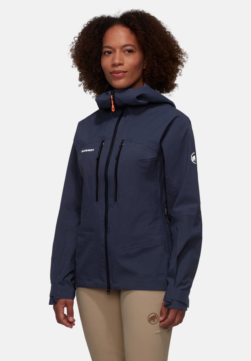 Marine buitenjas met capuchon, voorzien van twee met rits sluitende borstzakken, zwarte ritsen en een oranje accent op de ritslip. Gladde textuur.