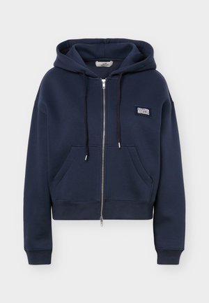 Tmavomodrá mikina na zips s prednými vreckami, kapucňou so šnúrkou a malou bielou logo nášivkou na ľavej strane hrude.