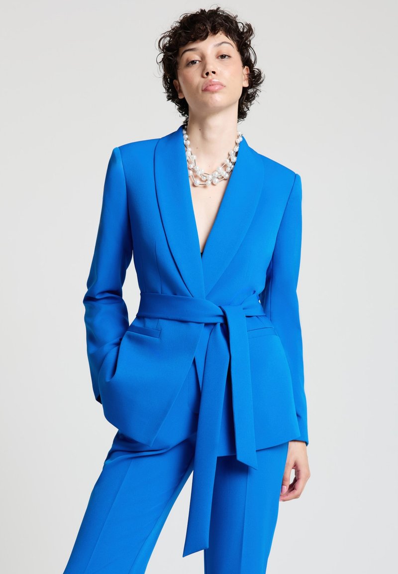 Woman wearing a vibrant blue belted suit with a deep V-neckline and a multi-strand pearl necklace, standing against a plain background.