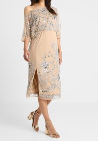 Beżowa sukienka do kolan z przezroczystego materiału z srebrnymi zdobieniami i kwiatowymi wzorami, z dekoltem typu off-the-shoulder oraz bocznym rozcięciem.