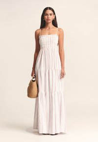 Vestido largo con corpiño fruncido, tirantes finos y falda de volantes; presenta tela blanca con rayas rojas. La modelo sostiene un bolso tejido.