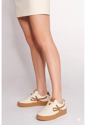 Piernas con falda beige y zapatillas blancas con rayas laterales marrones y suelas gruesas marrones sobre un fondo claro liso.