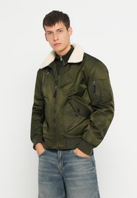 Veste bomber vert olive avec un col en fausse fourrure blanche, poches zippées, poignets côtelés et une finition légèrement brillante. Portée par-dessus un haut sombre.