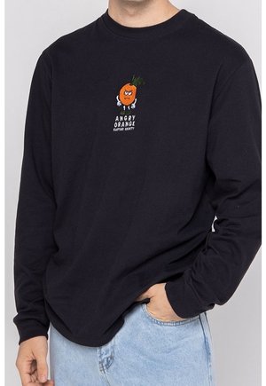 Sort langærmet skjorte med en vred orange tegneserie og teksten "ANGRY ORANGE KANTIKO SOCIETY", båret af en person med den ene hånd i lommen på lyseblå jeans.