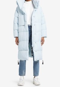 Ljusblå vadderad lång jacka med huva, med en frontdragkedja och en bröstficka. Buren över en vit topp och ljusblå jeans.