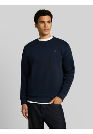 Mann trägt dunkelblauen Pullover über weißem Hemd und dunklen Jeans, steht vor schlichten hellgrauem Hintergrund und schaut nach rechts.