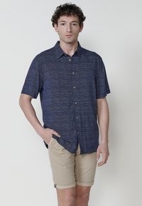 Camicia blu navy a maniche corte con motivo testurizzato, chiusura a bottoni e una vestibilità rilassata; abbinata a pantaloni corti beige chiaro con orli risvoltati.