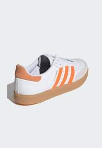 Białe sneakersy z pomarańczowymi paskami, zamszowym akcentem na pięcie, perforowanym noskiem i gumową podeszwą, zaprojektowane do codziennego użytku.