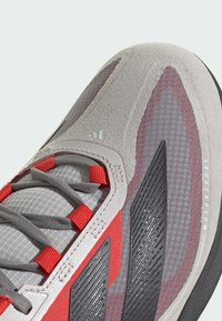 Close-up van een grijze en rode sneaker met zwarte, gestructureerde strepen, meshpanelen, rode lussen op de veters en de tekst "MOTORSPORT" op de rand van de zool.