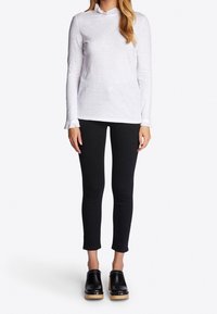 Vit långärmad topp med volangfåll runt halsen, kombinerad med svarta åtsittande byxor och svarta träskor med träliknande sula och metallaccenter.