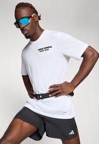 Bijela majica kratkih rukava s crnim natpisom "ADIDAS RUNNING SINCE 1949", u kombinaciji s crnim šortsevima koji imaju bijeli Adidas logo i crni pojas.