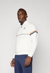 Hvit jakke med glidelås og ståkrage; har marineblå og oransje horisontale striper på ermene; myk stoff og figurnær design.