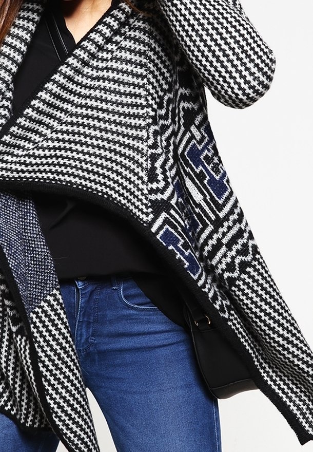 Personne portant un cardigan en tricot à motifs noir et blanc sur un haut noir et un jean bleu, avec un sac noir partiellement visible.