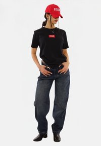 Black t-shirt with a red logo patch, loose blue denim jeans, and a red baseball cap featuring white text. Black ankle boots complete the look.